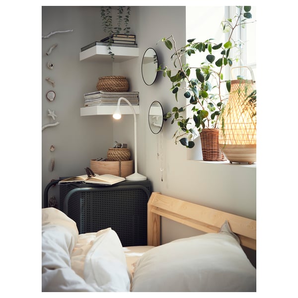 Ein gemütliches Schlafzimmer mit einer weißen Lampe auf einem Nachttisch neben dem Bett, einem hölzernen Kopfteil und einem Regal mit Büchern und Pflanzen.