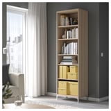 Hohes Bücherregal aus Holz mit mehreren Regalen, gefüllt mit Büchern und gelben Aufbewahrungsboxen. Positioniert in der Nähe eines Fensters mit durchsichtigen Vorhängen.