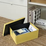 Eine gelbe NE-Schachtel auf einem Juteteppich, offen mit einem Buch und Papier darin. Regale mit Büchern dahinter.