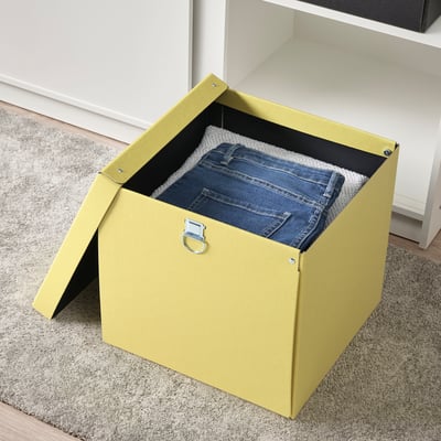 Eine gelbe NIMM Aufbewahrungsbox mit offenem Deckel, in der sich eine gefaltete Blue Jeans befindet. Die Box steht auf einem beigen Teppich in der Nähe eines weißen Schranks.