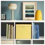 Bücherregal mit Büchern, einer gelben Box und Dekoration. Platte: Lampe, Kunst, gelbe Vase. Unten: bunte Bücher, gelbe Box, blaue Bücher.