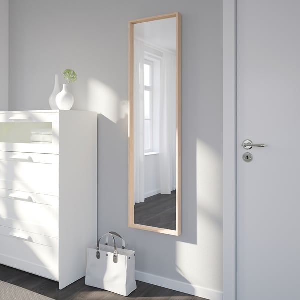 Großer Spiegel an der Wand, der den Raum reflektiert, zwischen weißer Kommode und Tür platziert. Tasche sitzt darunter, Vase mit Grün auf Kommode.