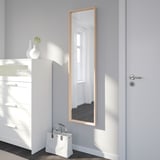 Großer Spiegel an der Wand, der den Raum reflektiert, zwischen weißer Kommode und Tür platziert. Tasche sitzt darunter, Vase mit Grün auf Kommode.