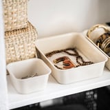 Weiße Aufbewahrungsboxen aus Kunststoff, eine quadratische, eine rechteckige, in denen Schmuck auf einem Regal aufbewahrt wird.