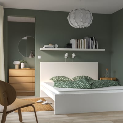 Modernes Schlafzimmer mit grünen Wänden, weißem Bett, Holzstuhl und Pendelleuchte. Das Bett hat grün gemusterte Kissen und befindet sich unter einem Regal mit Büchern.
