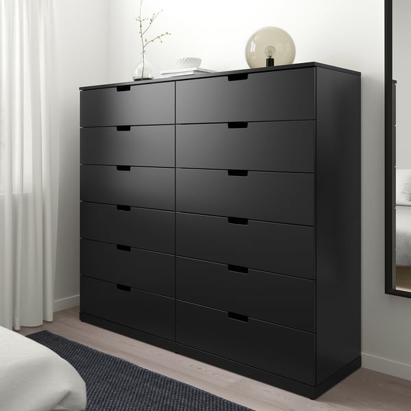 Alttext: schwarze NORDLI Kommode mit 9 Schubladen im Schlafzimmer, modern und hoch, mit hellen Verzierungen oben.