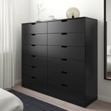 Alttext: schwarze NORDLI Kommode mit 9 Schubladen im Schlafzimmer, modern und hoch, mit hellen Verzierungen oben.