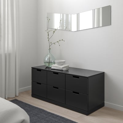 Moderne schwarze NORDLI-Kommode mit vier Schubladen, Spiegel oben, Vase, Bücher, Schale. Graue Wand, durchsichtige Vorhänge in der Nähe.