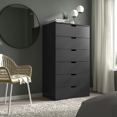 Die moderne graue NORDLI-Kommode steht hoch mit fünf Schubladen, in der Nähe eines Korbstuhls und eines großen Spiegels. Es ist ein schlankes, rechteckiges Design, das zur Schlafzimmereinrichtung passt.