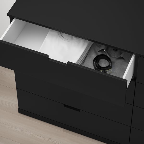 Schwarze Kommode NORDLI mit einer offenen Schublade, die den Blick auf die Kleidung freigibt, und einem Gürtel, der seine Aufbewahrungsfunktion zeigt.