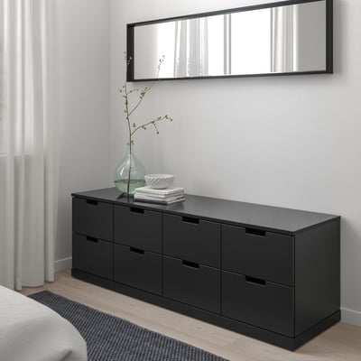 Schwarze NORDLI Kommode mit 6 Schubladen im Schlafzimmer, Spiegel, Fenster mit durchsichtigen Vorhängen, Vase mit Ästen.