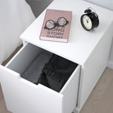 Weißer Nachttisch mit offener Schublade mit Decke, daneben Buch und Wecker.