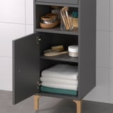 Hoher grauer Badezimmerschrank mit offenem und geschlossenem Stauraum. Bietet Platz für Handtücher, Toilettenartikel und Pflegewerkzeuge. Muss aus Sicherheitsgründen an der Wand montiert werden.