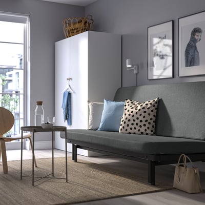 Modernes Wohnzimmer mit grauem NYHAMN-Schlafsofa, blauen Kissen, weißem Schrank und Wandkunst.