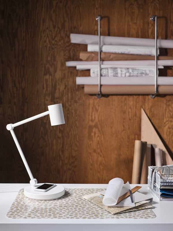 Moderne weiße verstellbare Schreibtischlampe mit kabellosem Ladegerät; Holzhintergrund, gemustertes Tischset, Papiere und Büromaterial.