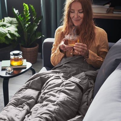 Die Frau entspannt sich mit einer ODONVIDE-Gewichtsdecke. Die Decke ist grau, weich und hat ein Box-Stitch-Muster. Sie trinkt Tee und sitzt bequem.