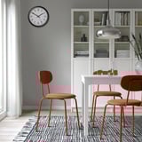 Drei orangefarbene ÖSTANÖ Stühle um weißen Tisch. Graue Uhr, Bücherregal mit Dekor. Kompakt, modern.