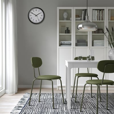 Moderne Esseinrichtung mit grünen Stühlen, weißem Tisch, silbernem Licht, gestreiftem Teppich, Uhr und Bücherregal.