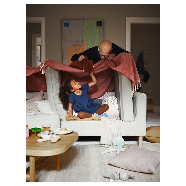Gemütliches Wohnzimmer mit Kind und Erwachsenem, die unter der Decke auf dem Sofa spielen. Modernes Sofa, Spielzeug, Snacks und Kissen sorgen für eine verspielte Atmosphäre.