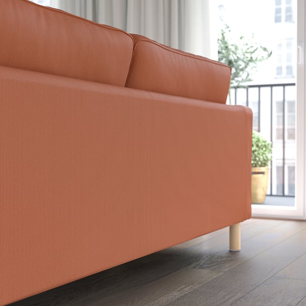 Orangefarbenes Sofa am Fenster mit klaren Linien und Holzbeinen. Topfpflanze auf dem Balkon draußen.