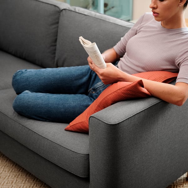 Eine Frau ruht sich auf einem grauen PÄRUP-Sofa aus und liest eine Zeitung. Das Sofa besteht aus einem abnehmbaren, maschinenwaschbaren Bezug und stützenden Taschenfederkern-Sitzkissen.