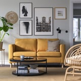 Modernes Wohnzimmer mit senffarbenem PÄRUP-Sofa, schwarzem Couchtisch, Wand aus Kunstgalerie und Korbstuhl.