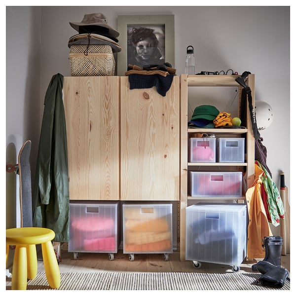 Holzschrank mit transparenten Kunststoffbehältern zur Aufbewahrung verschiedener Gegenstände. Enthält einen gelben Hocker, einen grünen Mantel und einen schwarzen Helm daneben.