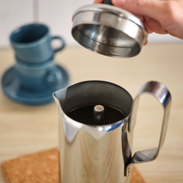 Die Hand hält den Metall-Kaffeetrichter über die silberglänzende PÅTÅR-Kaffeemaschine auf dem Tisch.