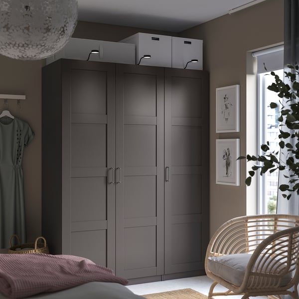 Modernes Schlafzimmer mit dunkelgrauem PAX-Kleiderschrank, hängendem Kleid, Wandkunst, Korbstuhl und großem Fenster.