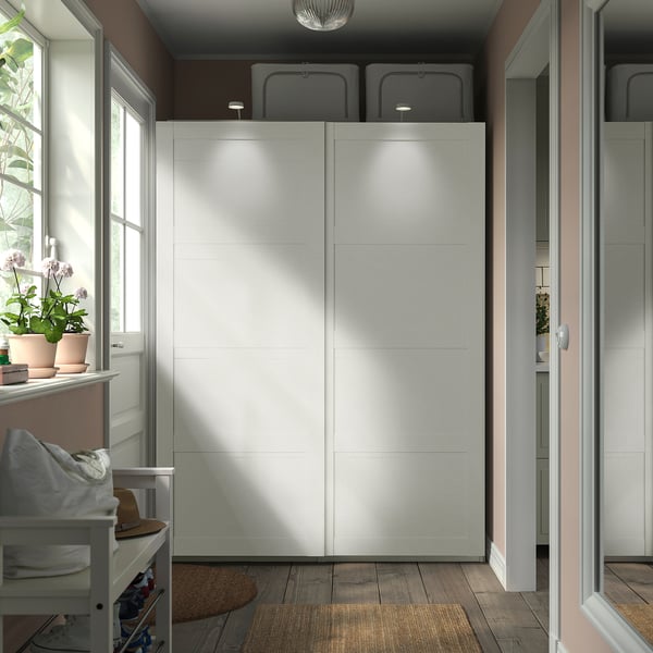 Großer weißer PAX-Kleiderschrank mit Schiebetüren, in der Nähe eines Fensters mit Topfpflanzen. Daneben steht ein kleiner Hocker mit zusammengefalteten Kleidern und Hut.