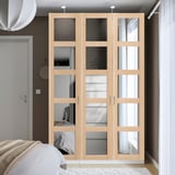 Ein hoher, heller PAX-Kleiderschrank mit verspiegelten Türen, der ein Bett und ein großes Fenster ergänzt. Es ist schlank und passt perfekt in die Ecke des Raumes.