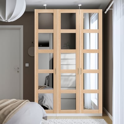 Ein hoher, heller PAX-Kleiderschrank mit verspiegelten Türen, der ein Bett und ein großes Fenster ergänzt. Es ist schlank und passt perfekt in die Ecke des Raumes.