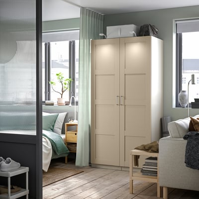 Modernes Schlafzimmer mit beigem PAX-Kleiderschrank mit integrierter Beleuchtung, hellem Holzboden, blau-grünen Vorhängen und gemütlicher Einrichtung.