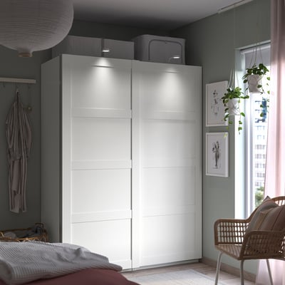 Modernes Schlafzimmer mit weißem PAX-Kleiderschrank, Schiebetüren, grauen Wänden, hängenden Pflanzen und einem Korbstuhl am Fenster.