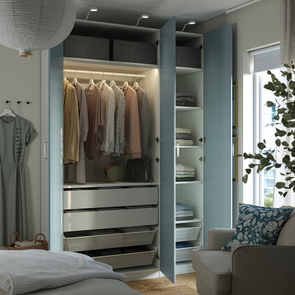 Schlafzimmer mit offenem blauem PAX-Kleiderschrank mit Kleidung, Schubladen und Regalen mit gefalteten Gegenständen, neben einem Bett und einem Stuhl.