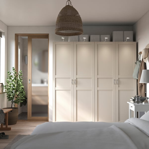 Modernes Schlafzimmer mit beigem PAX-Kleiderschrank, grauem Teppich, Pendelleuchte aus Korbgeflecht, weißem Bett, Holzboden und Zimmerpflanze.