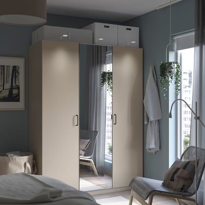 Modernes Schlafzimmer mit einem beigen PAX-Kleiderschrank, verspiegelten Türen und grauen Wänden. Eine Hängepflanze, ein Stuhl mit einer Decke und ein Bett, das teilweise sichtbar ist.
