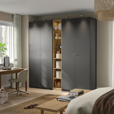 Modernes Schlafzimmer mit großem grauem PAX-Kleiderschrank und offenen Regalen, Schreibtisch und Stuhl, mit natürlichem Licht durch ein Fenster mit weißen Vorhängen.