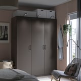 Modernes Schlafzimmer mit dunkelgrauem PAX-Kleiderschrank, weißen Boxen, hängendem Kleidungsstück, Pflanze, Stuhl und Fenster.