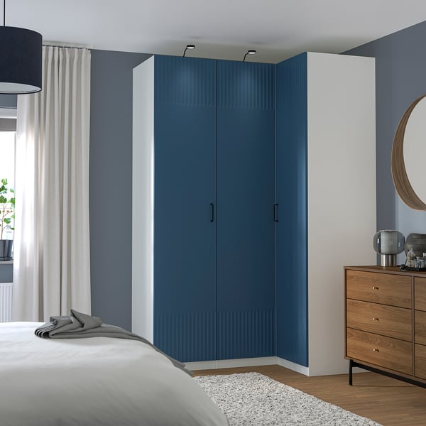 Großer blauer PAX-Kleiderschrank mit weißen Rahmen, modernem Design, steht hoch neben Holzschubladen und Bett, schlanke Griffe, vertikale Streifen.