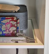 Ein weißer PAX-Kleiderschrank mit durchsichtigem Regal, in dem sich eine schwarze Hutschachtel befindet, und eine bunte perlenbesetzte Handtasche mit einem goldenen Kettenriemen.