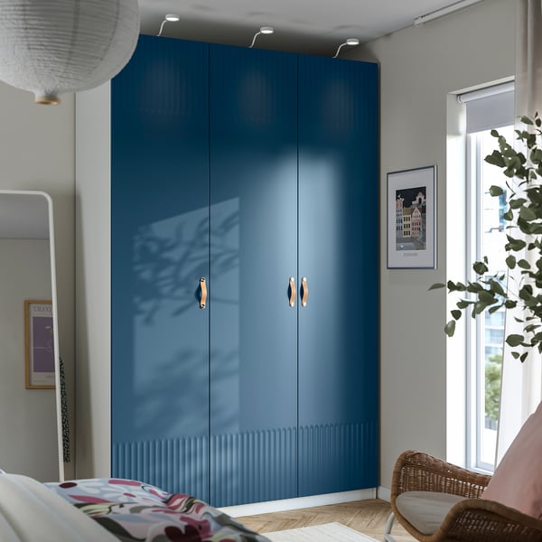 Ein moderner blauer PAX-Kleiderschrank mit schlanken Griffen und verspiegelten Türen. Er steht hoch neben einem Fenster, größer als eine Pflanze und ein Korbstuhl in der Nähe.