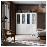 Modernes Schlafzimmer mit weißem PAX-Kleiderschrank mit Kleidung, Korbstuhl, gestreiftem Teppich, Holzboden und Pendelleuchte.