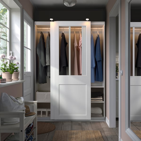 Ein weißer PAX-Kleiderschrank mit Glasschiebetüren, in denen die Kleidung ausgestellt ist. Er ist größer als andere Gegenstände im Raum, wie der weiße Hocker und die Blumentöpfe.