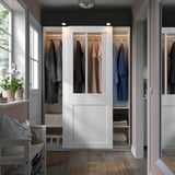 Ein weißer PAX-Kleiderschrank mit Glasschiebetüren, in denen die Kleidung ausgestellt ist. Er ist größer als andere Gegenstände im Raum, wie der weiße Hocker und die Blumentöpfe.