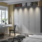 Grauer PAX-Kleiderschrank neben Schreibtisch und Fenster, mit schwarzem Stuhl, Lampe, Laptop. Das Zimmer verfügt über Pflanzen, hellgelbe Wände, Holzbalken und einen gemusterten Teppich.