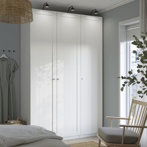 Modernes Schlafzimmer mit großem weißem PAX-Kleiderschrank, hoch und breit, neben einem Fenster mit Stuhl und Pflanze. Kleiderschrank mit mehreren Türen und minimalistischem Design.
