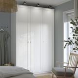 Modernes Schlafzimmer mit großem weißem PAX-Kleiderschrank, hoch und breit, neben einem Fenster mit Stuhl und Pflanze. Kleiderschrank mit mehreren Türen und minimalistischem Design.