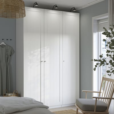 Modernes Schlafzimmer mit großem weißem PAX-Kleiderschrank, hoch und breit, neben einem Fenster mit Stuhl und Pflanze. Kleiderschrank mit mehreren Türen und minimalistischem Design.
