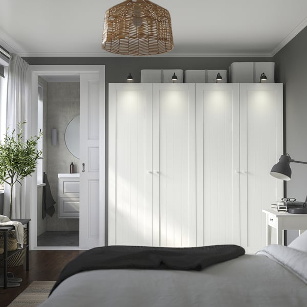 Modernes Schlafzimmer mit weißem PAX-Kleiderschrank, grauen Wänden, dunklem Boden und Lampe. Blaues Bett im Vordergrund, Badezimmertür offen.
